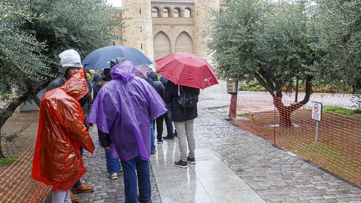 Varias personas se refugian con chubasqueros y paraguas de la lluvia en La Aljafería de Zaragoza
