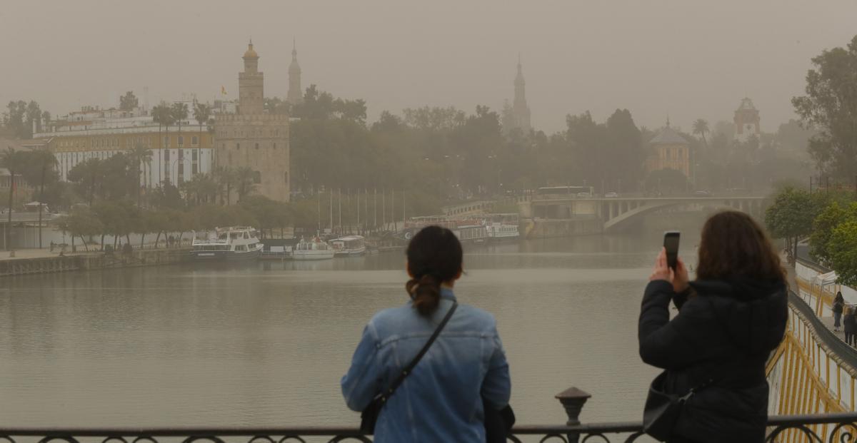 Dos jóvenes fotografían la Torre del Oro de Sevilla con el cielo cubierto por la intensa calima debido al polvo procedente del desierto del Sáhara.