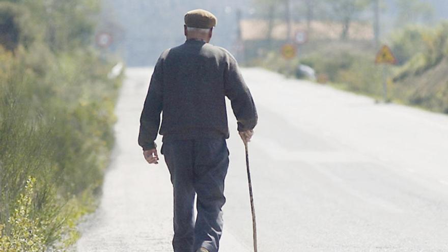 Un anciano camina por una carretera gallega apoyado en un bastón.