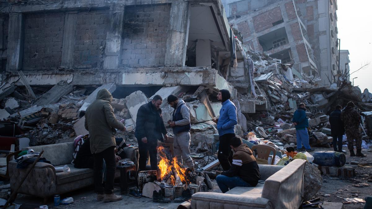 Varias personas calentándose alrededor del fuego y entre los escombros de un edificio derrumbado.