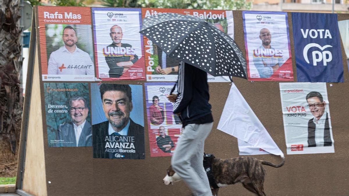 Un vecino de Alicante pasando con su paraguas por delante de un panel con carteles electorales durante este temporal de lluvias.