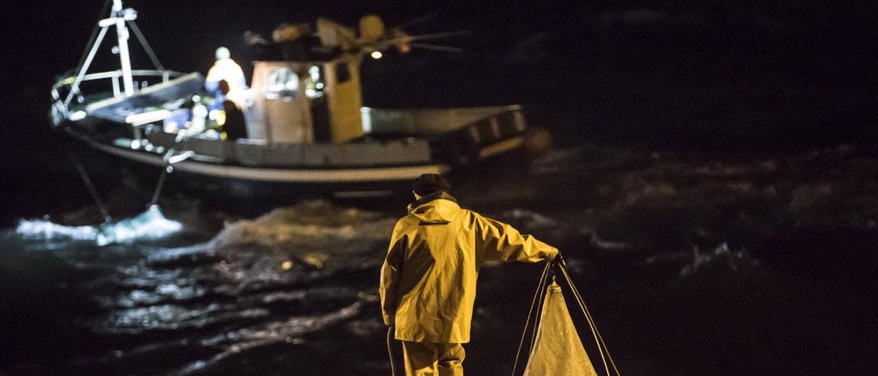 Un angulero a pie mira cómo trabajan pescadores a bordo de una lancha.