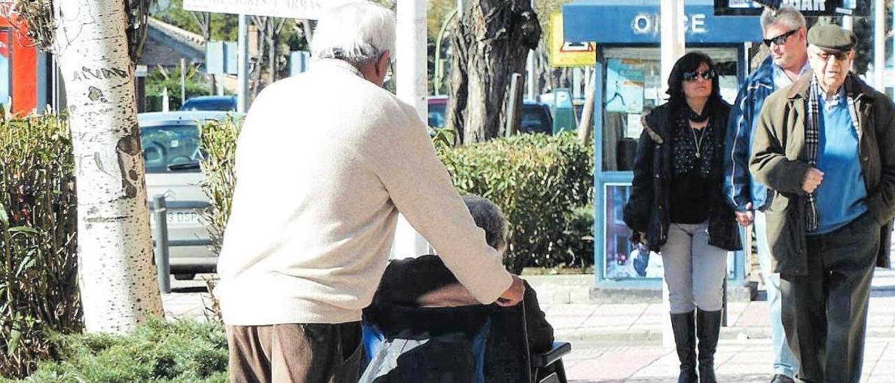 Una persona dependiente en silla de ruedas y su acompañante pasean por la calle.