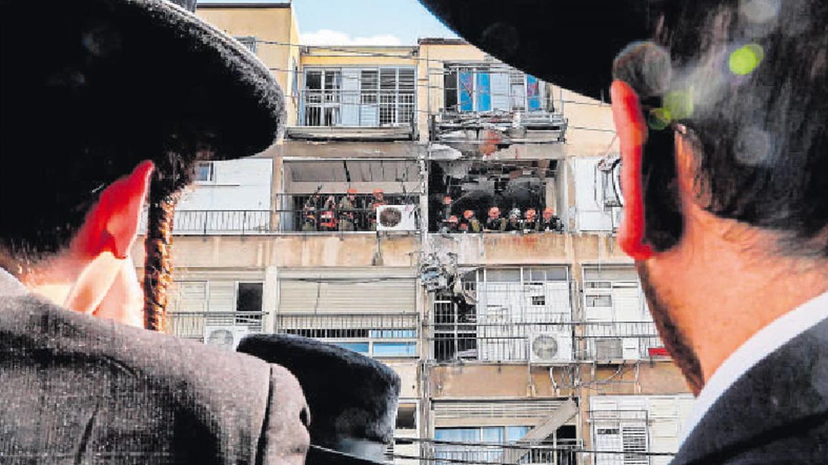 Jueus ultraortodoxos observen els militars israelians durant la inspecció de l’edifici bombardejat per l’Exèrcit de l’Iran al barri residencial Bnei Brak, a Tel-Aviv, ahir.