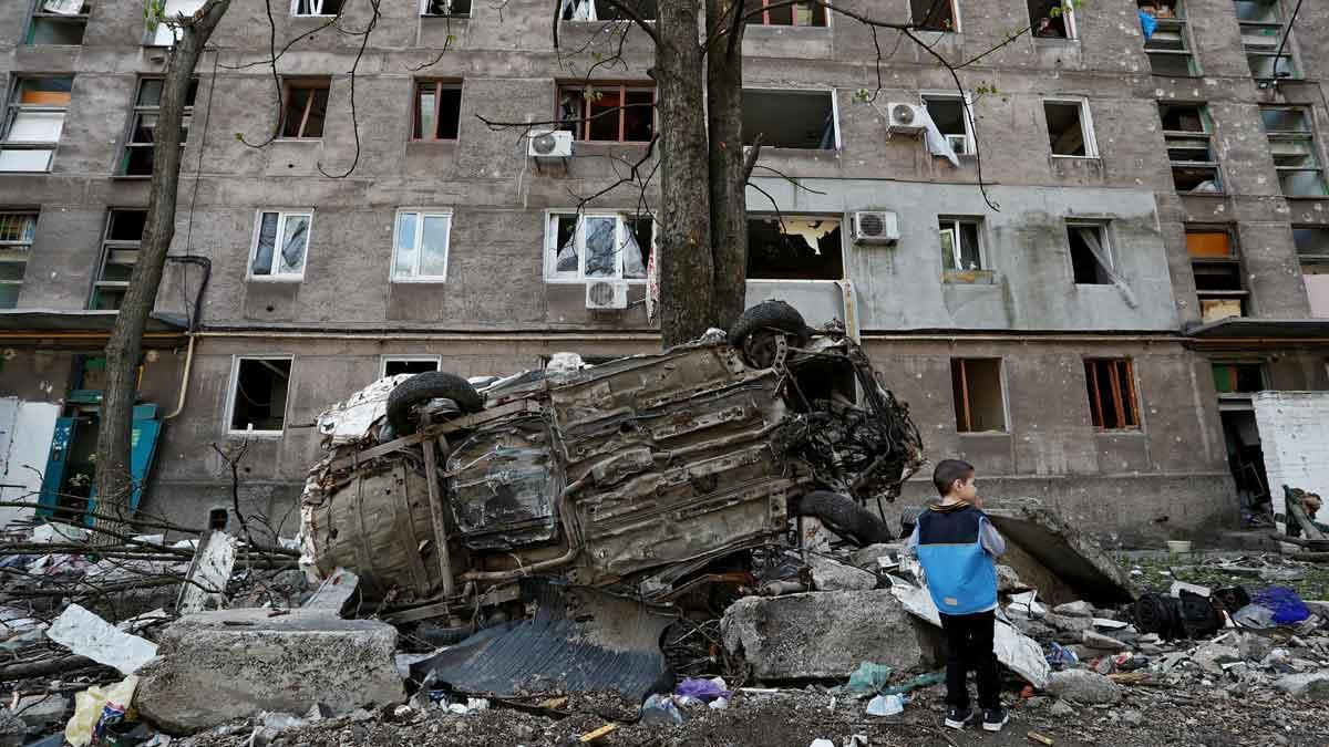 Un niño junto a un coche destrozado y un edificio de viviendas dañado por los ataques en la ciudad de Mariúpol