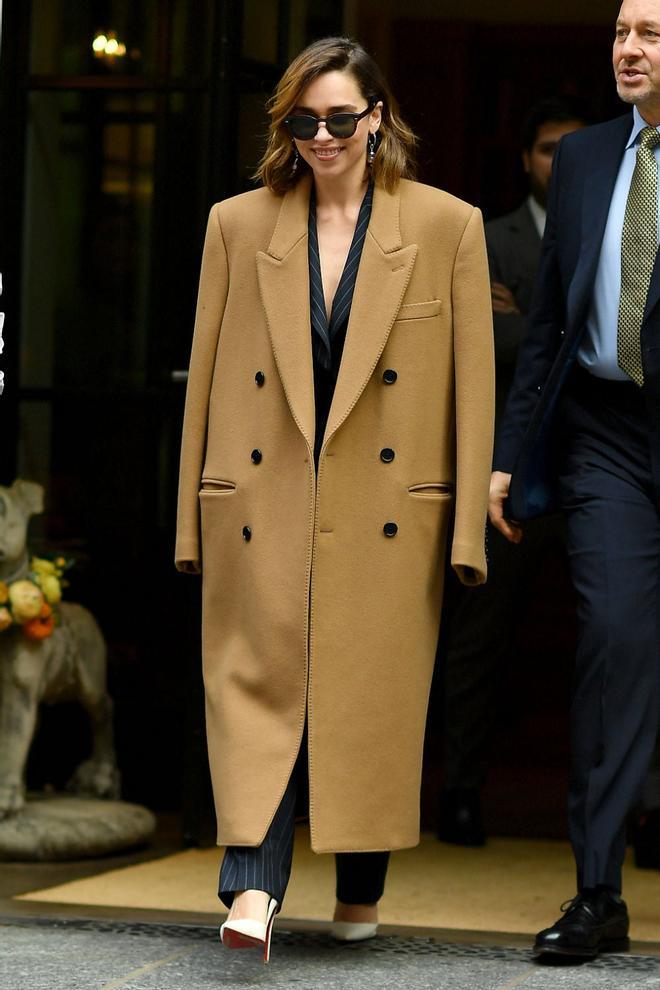 Emilia Clarke combina un traje de chaqueta y pantalón de raya diplomática con un abrigo camel oversized de aire masculino y unos salones blancos