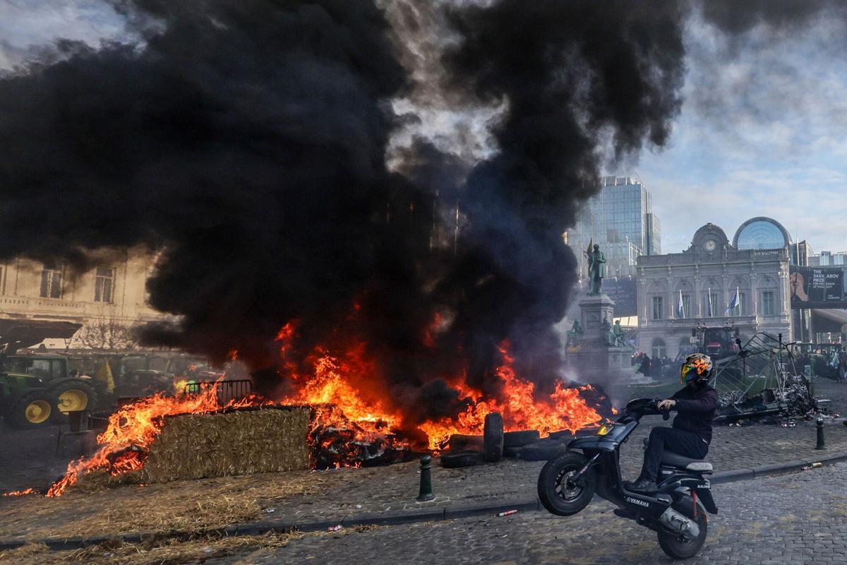 Un motociclista realiza una maniobra frente a una pila de neumáticos en llamas durante la masiva protesta agraria en Bruselas. Miles de productores europeos se han manifestado ante las instituciones de la Unión para rechazar el acuerdo con Mercosur, las reformas de la PAC y la creciente presión regulatoria. 18 de diciembre de 2025, Bruselas.