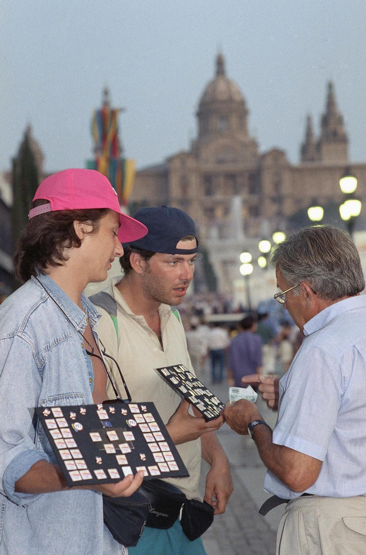 Vendedores de pins olímpicos en la avenida de la Reina María Cristina de Barcelona. Jeff Morgan (izquierda), de Granada Hills, California, y David Langenthrow,  de Oakland, California, negocian el precio de un pin con Paul Lewis, de Austin, Texas, el 6 de agosto de 1992. Durante los Juegos Olímpicos, los pins se conviertieron casi en moneda de cambio internacional y se vendían y cambiaban por dinero, comida, bebida, transporte e incluso periódicos.