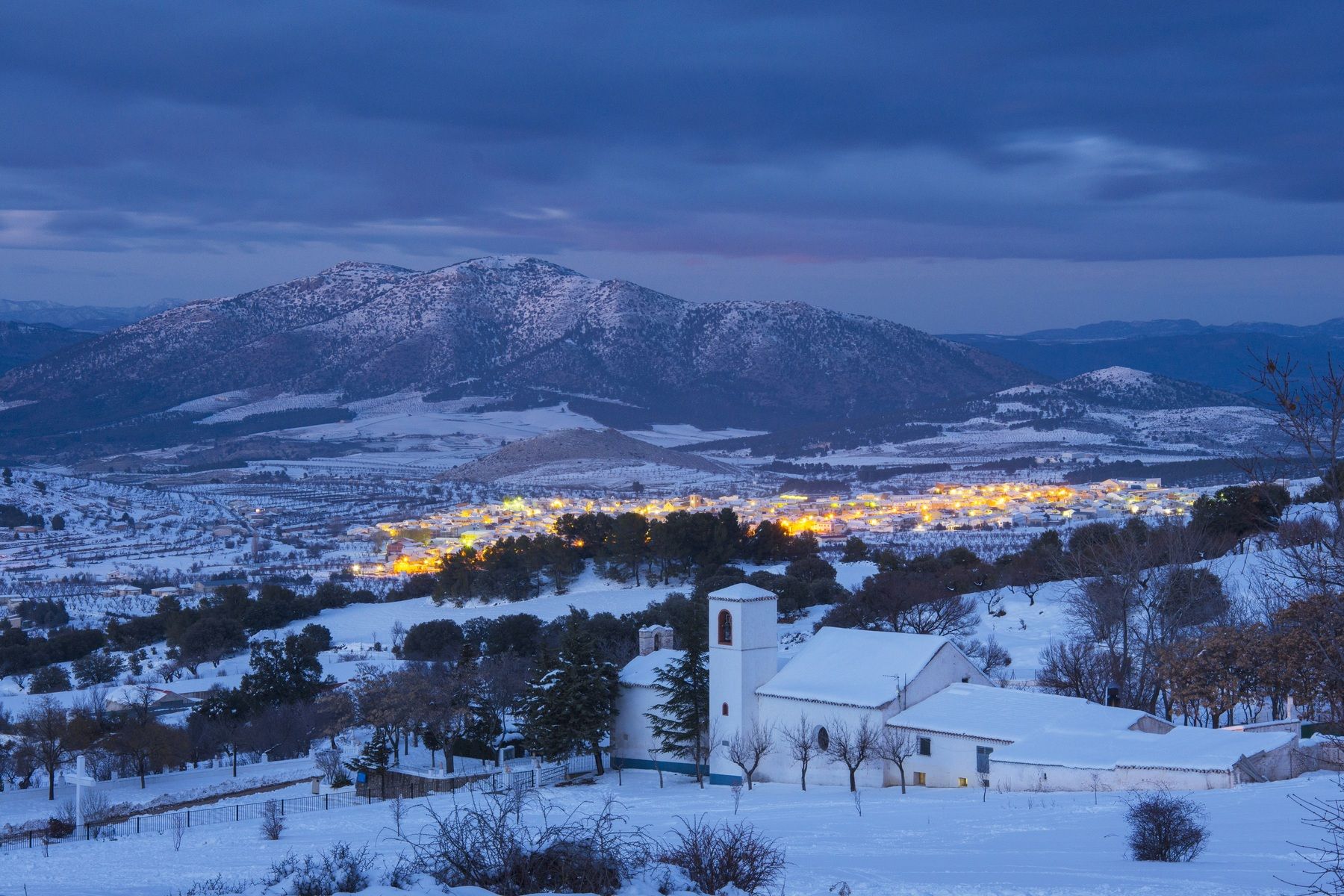 Vista invernal de María, en la comarca de Los Vélez (Almería), con el casco urbano iluminado y la Sierra de María-Los Vélez cubierta de nieve al fondo.