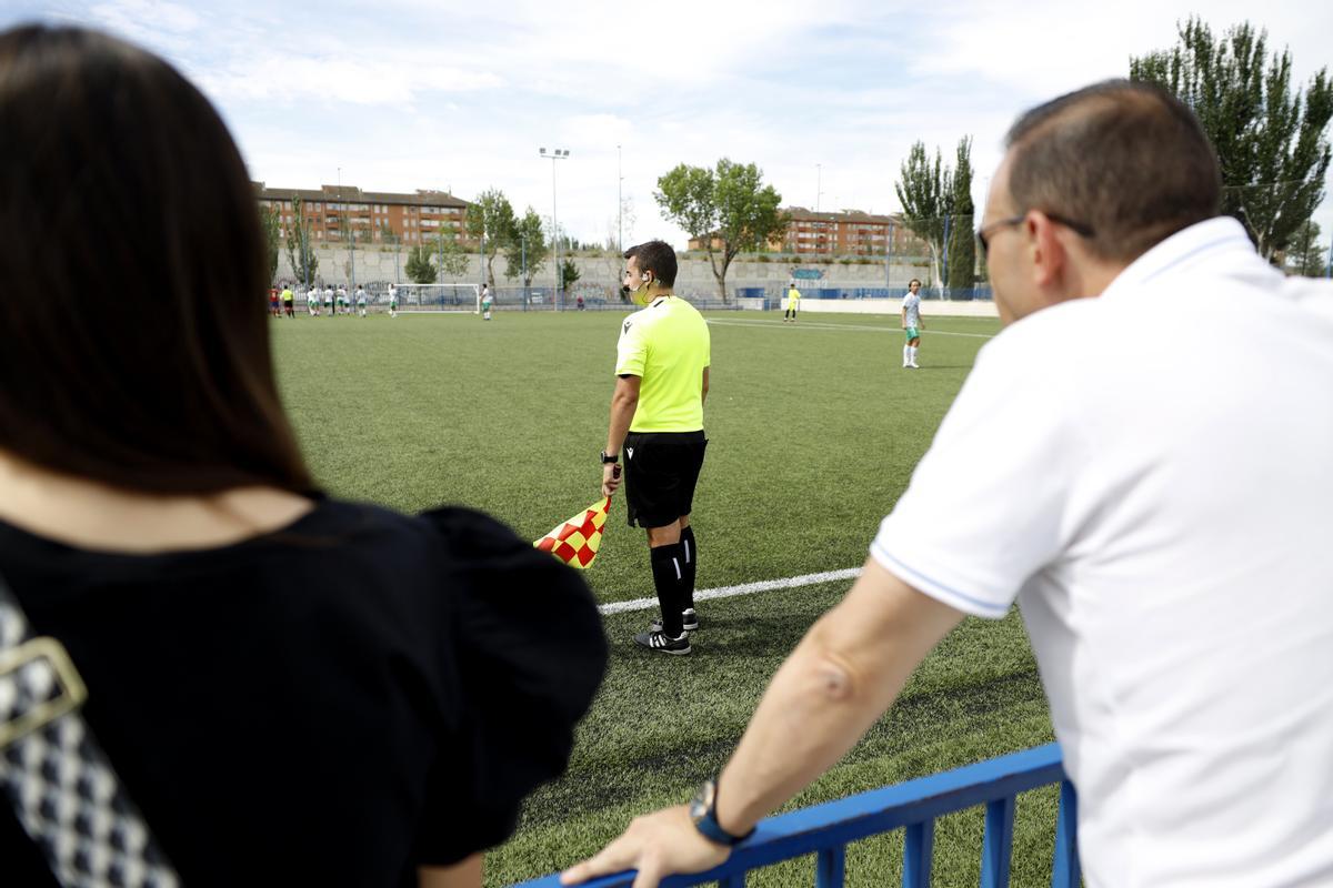 Dos espectadores presencian un partido de fútbol base en Aragón junto al juez de línea del mismo.