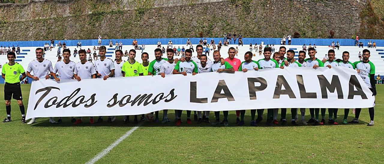 Los dos equipos y el trío arbitral, con la pancarta ante del inicio del partido.