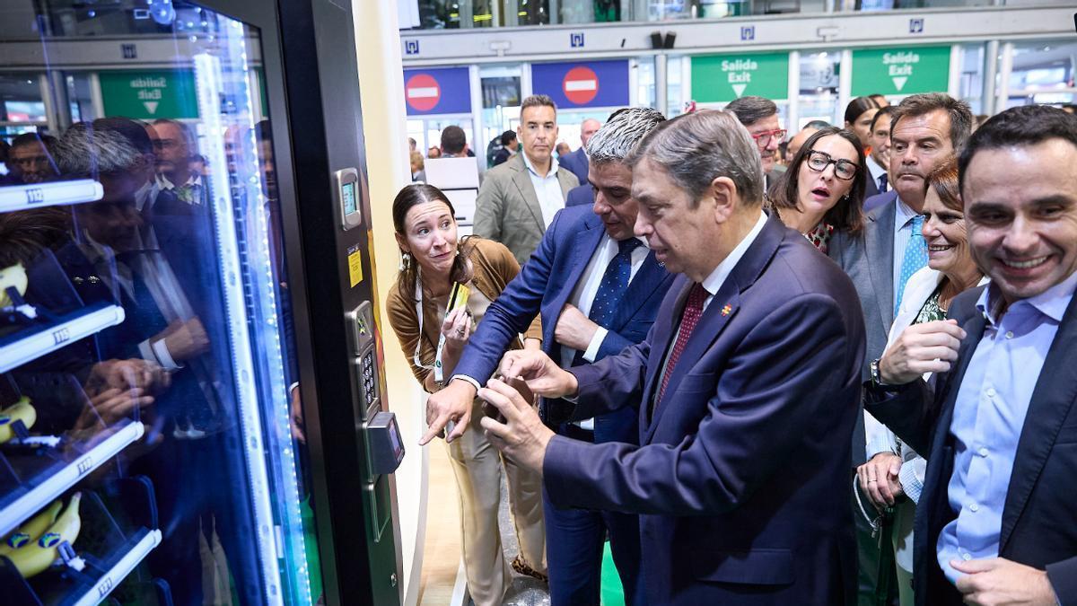 El consejero de Agricultura, Narvay Quintero, ayuda al ministro Luis Planas a comprar un plátano canario en una máquina expendedora.