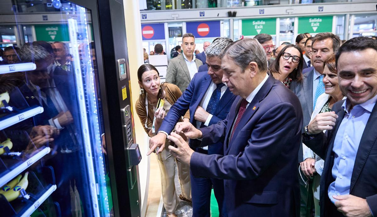 El consejero de Agricultura, Narvay Quintero, ayuda al ministro Luis Planas a comprar un plátano canario en una máquina expendedora.