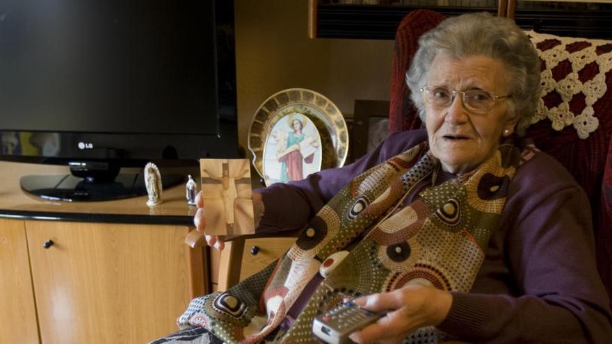 Pepica Alfaro, de 84 años, sostiene una estampa y el mando del televisor en su casa de Xàtiva, donde sigue cada domingo la misa.