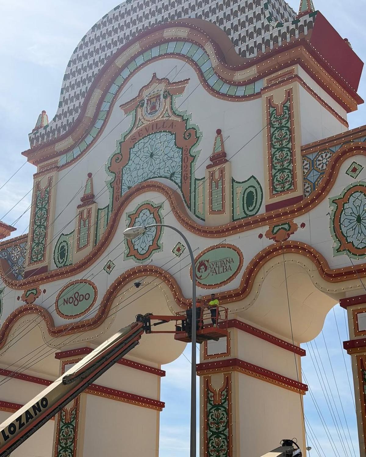 Una grua con dos operarios colocan el recuerdo a la duquesa de Alba en la Portada de la Feria de Abril de Sevilla.