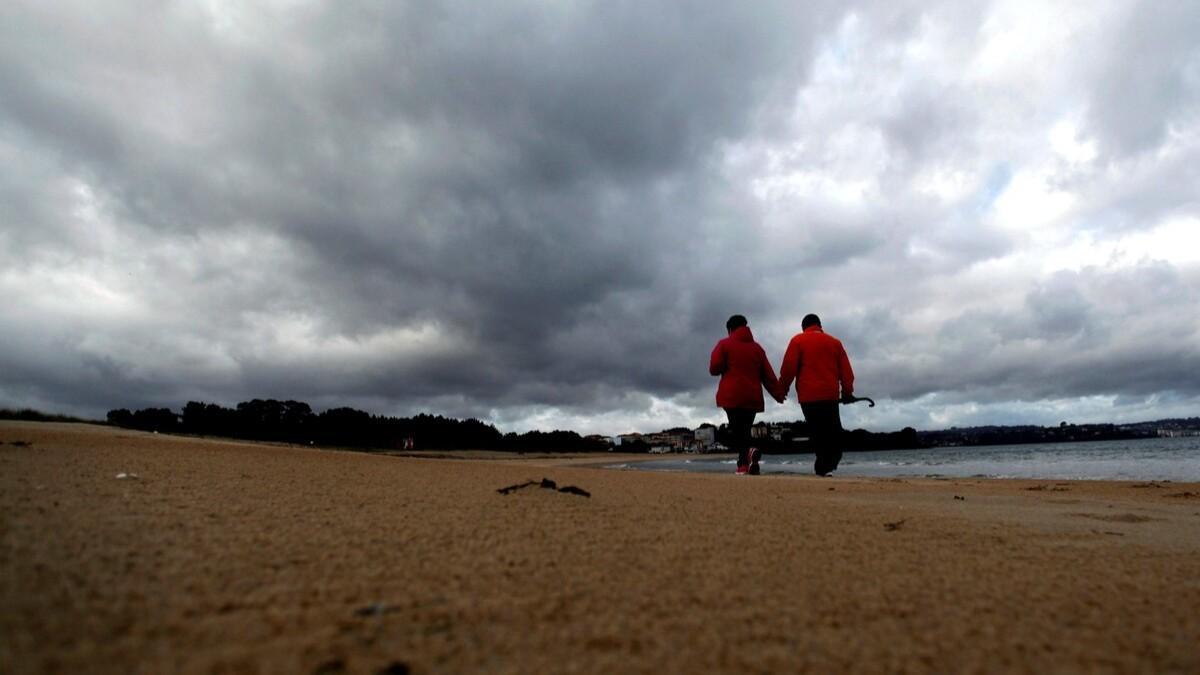 Una pareja pasea por una playa en Galicia un día con el cielo cubierto por las nubes