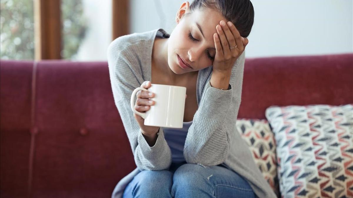 FOTO BAJADA DE 123RF EL 14 DE ENERO DEL 2021 PARA ILUSTRAR UN ARTICULO SOBRE EL  BLUE MONDAY   EL DIA MAS TRISTE DEL ANO SEGUN UN ALGORITMO MATEMATICO  CHICA TRISTE  TRISTEZA  DEPRESION  REFLEXION  shot of depressed young woman having headache while drinking coffee on sofa at home  FOTO DE 123RF