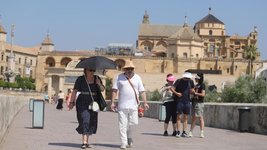 Varias personas se protegen del sol con gorras, sombreros y paraguas mientras caminan por el Puente Romano de Córdoba.