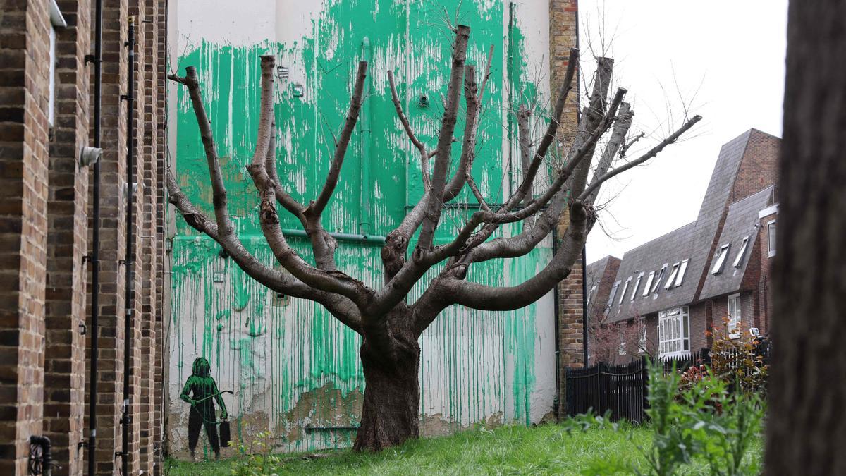 Obra de arte de Banksy, una plantilla de una persona que ha pintado con aerosol el follaje de un árbol en una pared detrás de un árbol sin hojas, cerca de Finsbury Park en el norte de Londres.