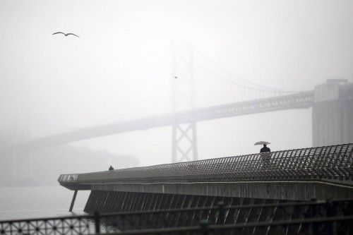 A man walks beneath an umbrella along Pier 14 near the San Francisco Oakland Bay Bridge during a light rain in San Francisco